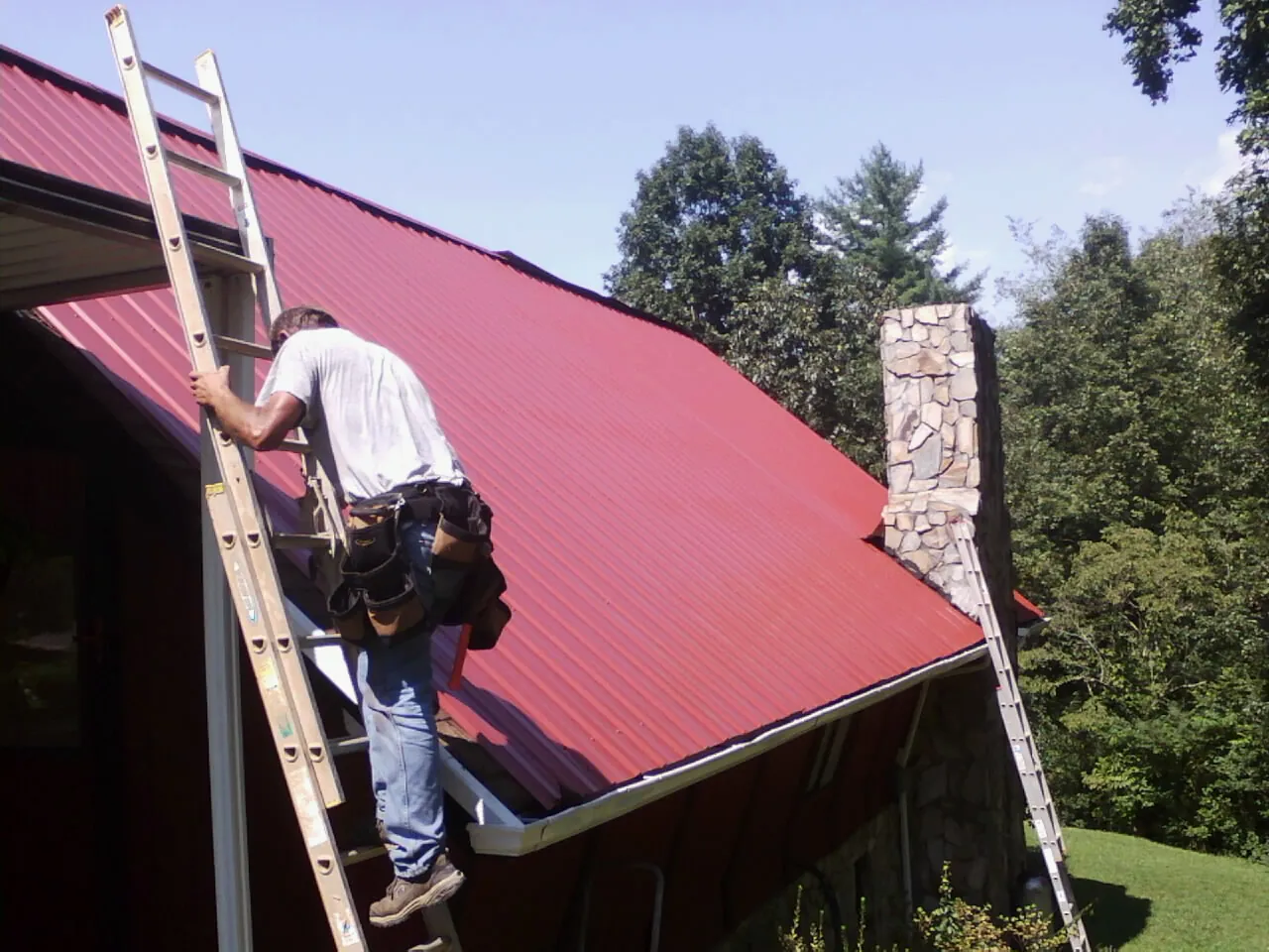 Roofer on a ladder beside a red metal roof and stone chimney — Soffit & Fascia Repair by Roofing Brothers in Bradford